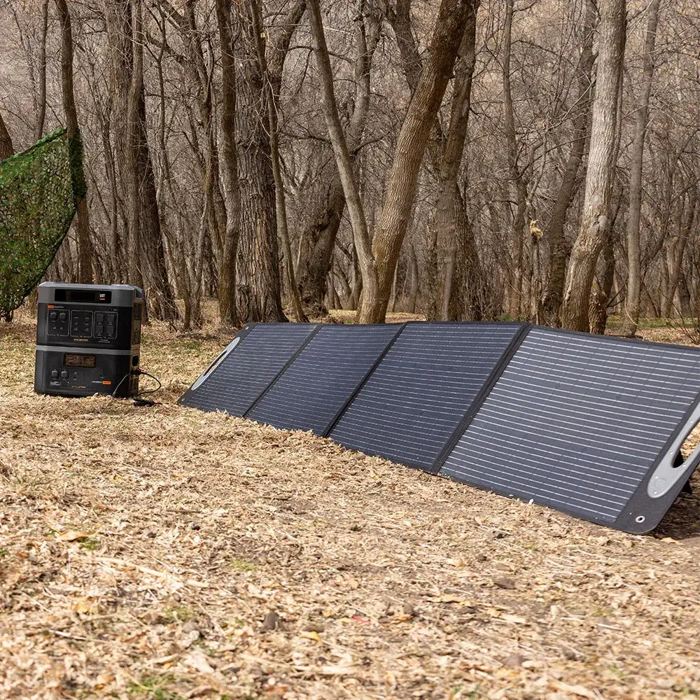 A Grid Doctor 3300 solar generator standing on a leaf-covered forest floor beside its folding solar panel array, angled toward the sun at a wooded campsite