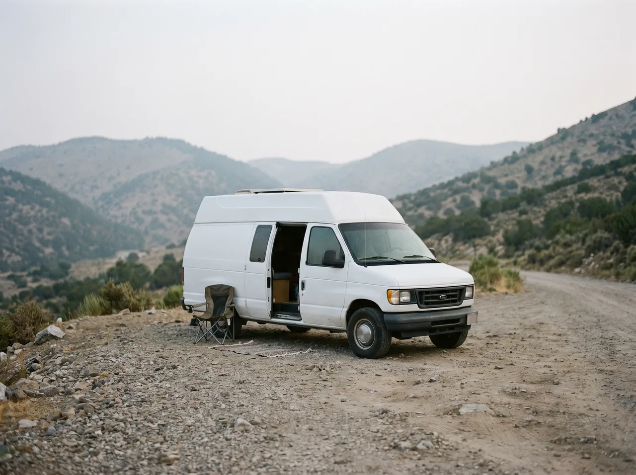 A camper van parked on a gravel pullout with its side door slid open, hills soft in the distance.